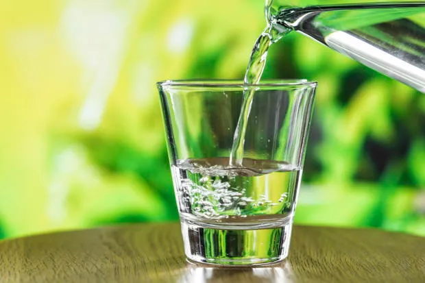Water being poured into a clear glass on a wooden surface with a green background.
